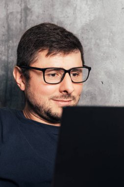 Modern ofisteki beton duvarın arkasında dizüstü bilgisayarına bakan gözlüklü yakışıklı sakallı adam. Yakın görünüm