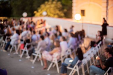 İnsanlar klasik müzik konseri izliyorlar. Bulanık genç ve yetişkin kadınlar, erkekler ve diğer insanlar klasik orkestranın konserini açık havada dinliyorlardı..