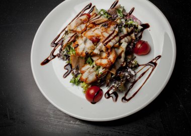 Kalamarlı salata, marul, lahana, vişneli domates ve sos. Kalamar dilimli salata, beyaz tabakta, siyah arka planda. Yakın plan fotoğraf, yumuşak odaklanma. Yemek tarifi kitabı için fotoğraf, restoran menüsü