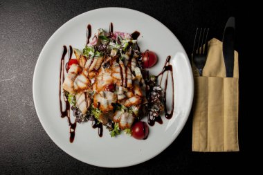 Kalamarlı salata, marul, lahana, vişneli domates ve sos. Kalamar dilimli salata, beyaz tabakta, siyah arka planda. Yakın plan fotoğraf, yumuşak odaklanma. Yemek tarifi kitabı için fotoğraf, restoran menüsü