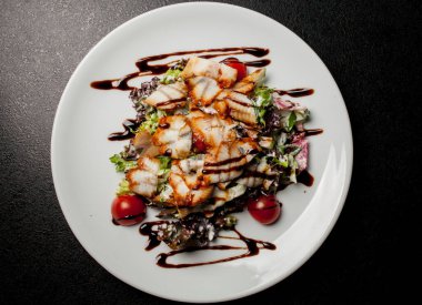 Kalamarlı salata, marul, lahana, vişneli domates ve sos. Kalamar dilimli salata, beyaz tabakta, siyah arka planda. Yakın plan fotoğraf, yumuşak odaklanma. Yemek tarifi kitabı için fotoğraf, restoran menüsü