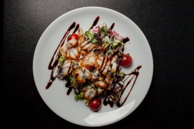 Kalamarlı salata, marul, lahana, vişneli domates ve sos. Kalamar dilimli salata, beyaz tabakta, siyah arka planda. Yakın plan fotoğraf, yumuşak odaklanma. Yemek tarifi kitabı için fotoğraf, restoran menüsü