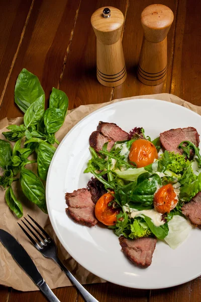 Meat beef steaks with greens, apricots and sauce on a large white plate on food mat - bamboo mat for food background. There are cutlery, a wooden pepper shaker and a salt shaker. barbeque, BBQ