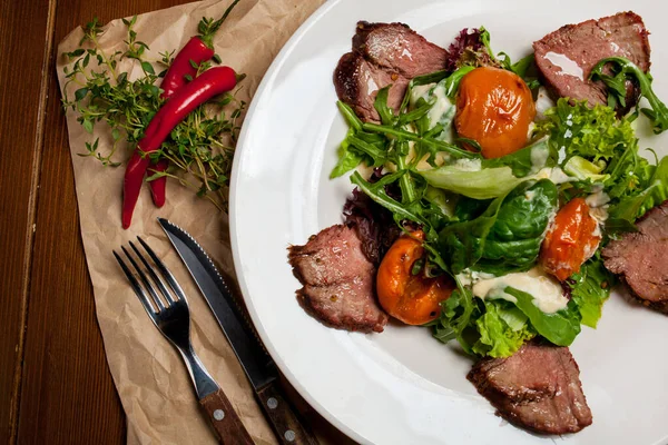 Meat beef steaks with greens, apricots and sauce on a large white plate on food mat - bamboo mat for food background. There are cutlery, a wooden pepper shaker and a salt shaker. barbeque, BBQ