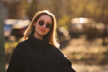 Siyah ceketli ve siyah güneş gözlüklü sarışın bir kız sokakta yürüyor. Siyah ceketli ve siyah gözlüklü güzel bir kız. yumuşak odak.