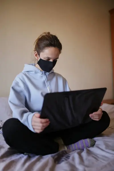 Mavi kapüşonlu ve siyah maskeli beyaz kız evde tek başına dizüstü bilgisayarda çalışıyor. Maskeli ve mavi kapüşonlu bir kız evde dizüstü bilgisayarda çalışıyor, yatakta oturuyor. Kendini soyutlama. Karantinaya alındı. COVID-19