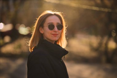 Siyah ceketli ve siyah güneş gözlüklü sarışın bir kız sokakta yürüyor. Siyah ceketli ve siyah gözlüklü güzel bir kız. yumuşak odak.