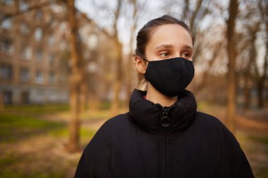 Siyah ceketli bir kız ve parkta siyah bir maske. Siyah tıbbi maskeli bir kız sokakta. Karantinaya alındı. Kendini soyutlama. Siyah tıbbi maskeli bir kızın portresi..
