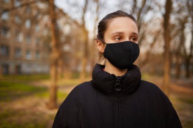 Siyah ceketli bir kız ve parkta siyah bir maske. Siyah tıbbi maskeli bir kız sokakta. Karantinaya alındı. Kendini soyutlama. Siyah tıbbi maskeli bir kızın portresi..