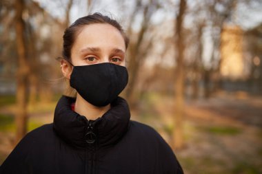 Siyah ceketli bir kız ve parkta siyah bir maske. Siyah tıbbi maskeli bir kız sokakta. Karantinaya alındı. Kendini soyutlama. Siyah tıbbi maskeli bir kızın portresi..