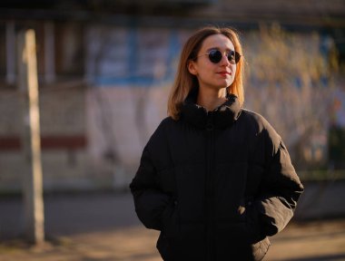 Siyah ceketli ve siyah güneş gözlüklü sarışın bir kız sokakta yürüyor. Siyah ceketli ve siyah gözlüklü güzel bir kız. yumuşak odak.