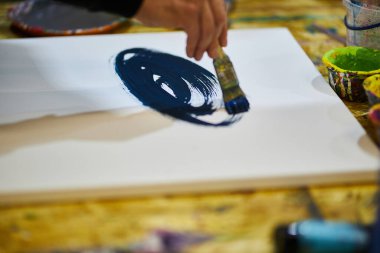 Bir adam beyaz bir tuvale siyah, mavi akrilik boya sürerek resim yapar..