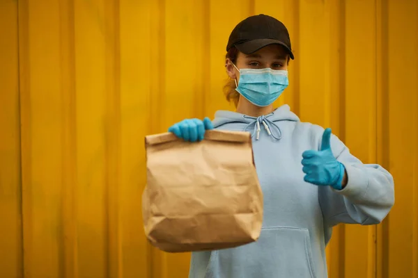 Tıbbi maske ve tıbbi eldiven takmış bir kız kurye. Karantinada teslimat servisi. Beyaz, tatlı bir kadın, teslimatçı, elinde bir market poşeti var. Kız kurye süper el hareketi gösteriyor.