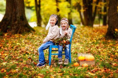 Bej kazaklı, kot pantolonlu ve şapkalı iki neşeli çocuk parkta bir sandalyede, balkabağının ve sonbahar sarısı yapraklarının yanında oturuyor.