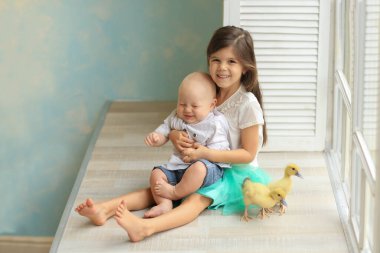 Beyaz tişörtlü ve mavi etekli kız beyaz gömlekli ve kot pantolonlu bebeğe sarılıyor pencerenin kenarında oturan iki yavru ördeğin yanında. Paskalya. Çocuklar ve hayvanlar