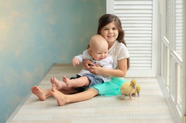 Beyaz tişörtlü ve mavi etekli kız beyaz gömlekli ve kot pantolonlu bebeğe sarılıyor pencerenin kenarında oturan iki yavru ördeğin yanında. Paskalya. Çocuklar ve hayvanlar