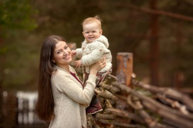 Kazak giymiş gülümseyen bir anne köydeki çitin üzerinde oturan küçük bir kızı tutuyor.
