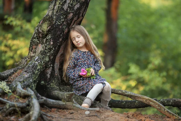 長い巻き毛のブロンドの髪の少女は木の下で夢を見て森の中に座って花を手に — ストック写真