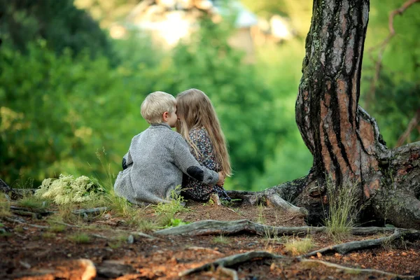 男孩穿着西服 扮演一个卷曲长发的女孩 穿着衣服坐在森林的树根上 — 图库照片