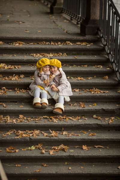 Маленькие сестры-близнецы в осенних пальто и желтых беретах обнимаются сидя на лестнице