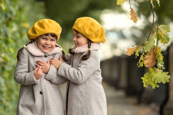 Zwei Kleine Zwillingsschwestern Herbstmantel Und Gelben Hüten Stehen Einer Eiche lizenzfreie Stockfotos
