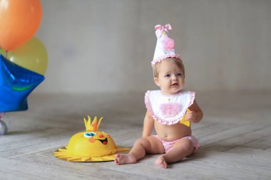 Pembe şapkalı ve önlüklü küçük kız balonların yanındaki tahta zeminde oturuyor ve doğum günü pastası yiyor. Doğum günü, yıl