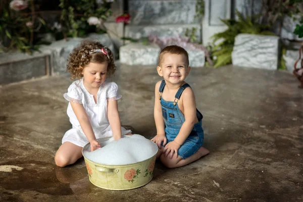 女の子で白いドレスにカールと彼女の笑顔弟でデニムショートパンツで洗面台で遊ぶでシャボン玉で夏の庭 — ストック写真