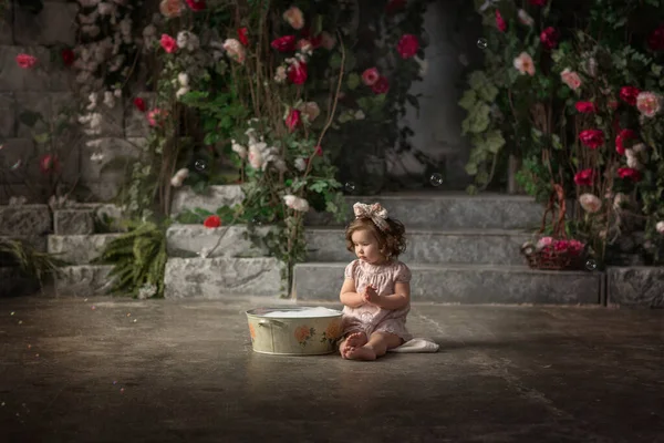 可愛い女の子がバラの茂みの庭に座ってシャボン玉を捕まえて — ストック写真