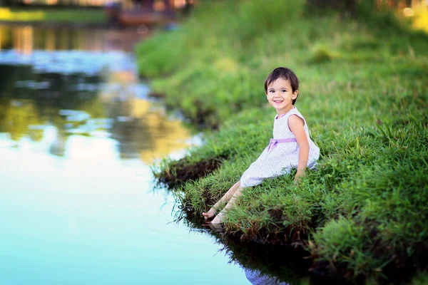 湖のそばに草の上に座っている裸の足を持つドレスのかなり小さな女の子 — ストック写真