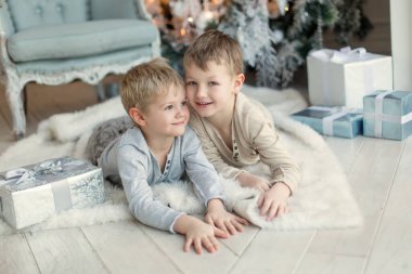 Noel ağacının yanında açık renk elbiseli iki küçük çocuk hediyeleri açıyor. Yeni Yıl