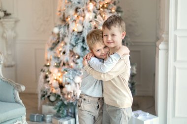 Noel ağacının yanında açık renk elbiseli iki küçük çocuk hediyeleri açıyor. Yeni Yıl