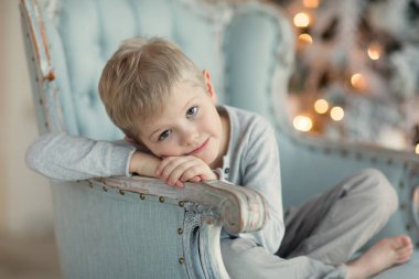 Sarışın bir çocuk, Noel ağacının yanındaki mavi koltukta oturmuş gülüyor.