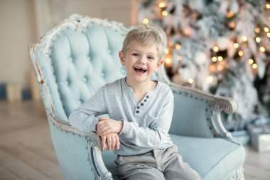 Sarışın bir çocuk, Noel ağacının yanındaki mavi koltukta oturmuş gülüyor.