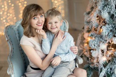 Güzel mavi gözlü bir anne ve genç bir oğlu Noel ağacının yanında sarılıyor. Yeni yıl için tebrikler. Aile tatili