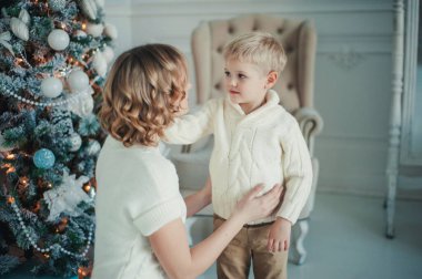 Beyaz örgü elbiseli güzel bir anne ve beyaz süveterli oğulları Noel ağacının yanında oturuyor.