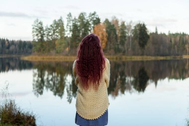 Sıcak kazak giyen genç bir kadın sonbaharda bir parkta gölün kenarında duruyor.