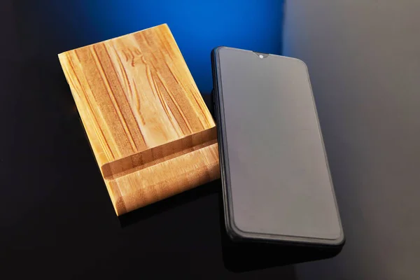 Wooden desktop mobile phone holder with hand-held phone isolated on blue black background. Creativity and technology concept.