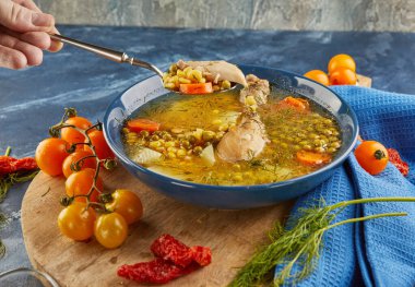 Tavuklu bezelye çorbası, havuç, domates ve otlar ahşap bir tabakta, mavi arka planda peçeteyle.