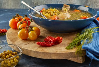 Tavuklu bezelye çorbası, havuç, domates ve otlar ahşap bir tabakta, mavi arka planda peçeteyle.