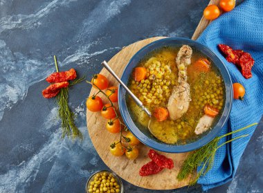 Tavuklu bezelye çorbası, havuç, domates ve otlar ahşap bir tabakta, mavi arka planda peçeteyle.