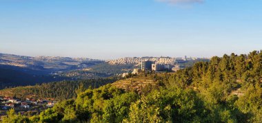 Parlak gün ışığında verimli orman tepelerinin üzerinde yükselen şehir silüeti yeşil doğal manzara boyunca uzaktan görülüyor..