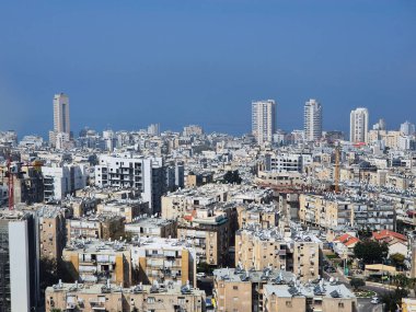 Mavi gökyüzünün altında deniz ufkuna kadar uzanan yoğun binaların bulunduğu şehir manzarası, şehir ve doğal elementler arasındaki zıtlığı vurguluyor..