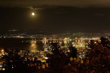 Haifa limanının üzerindeki ay ışığı denizi yansıtıyor, sanayi vinçleri ve şehrin ışıkları rıhtımda parlıyor..