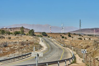 Kuru tarlalar ve elektrik hatlarıyla çevrili rüzgar türbinlerine ve dağ sırtlarına doğru giden dolambaçlı kırsal bir yol..