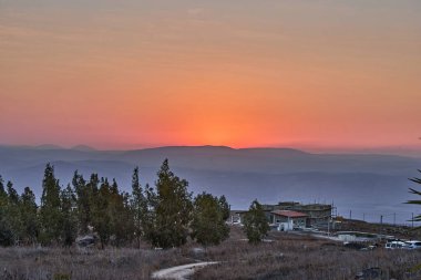 Gün batımı, dağların zirvelerine ulaşırken katmanlı tepelerin üzerinde parlayan turuncu ufuklar ve sakin kırsal kesimde siluetli ağaçlar görülür..