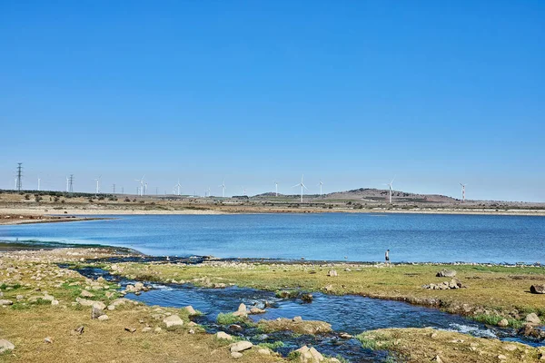 Mavi göl kayalık kıyı şeridi ve yeşil yosunlarla çevrili. Rüzgar türbinleri ve açık gökyüzü var..