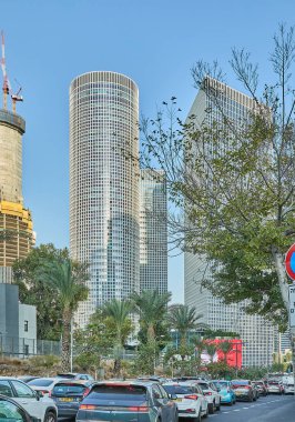 Tel Aviv, İsrail - 19 Kasım 2025: Park halindeki arabaların, yapraklı ağaçların ve Tel Aviv gökyüzünün altında yükselen ikonik yuvarlak kulelerin olduğu şehir trafik sahnesi.
