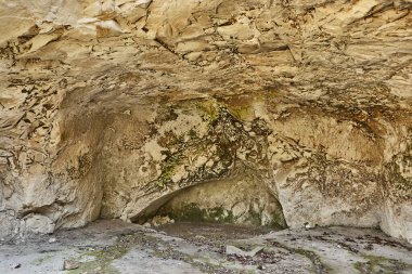 Kaya duvarları, yosun parçaları ve toprak zemin içeren doğal bir mağaranın içinde jeolojik oluşumlar ve doğal ışıklandırmaları vurguluyor.