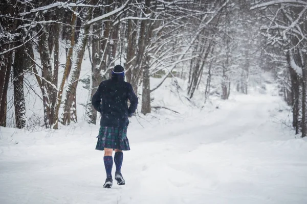 Man in kilt jogging in winter forest road while snowing