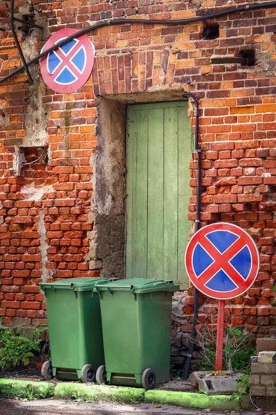 Two garbage bins standing on street with red brick building wall with green wooden door and two restrictive road signs